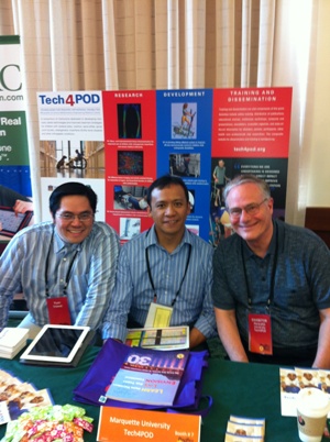Three men sit at a booth for Marquette University Tech4POD in front of an informational poster about Tech4POD and pamplets on the table. 